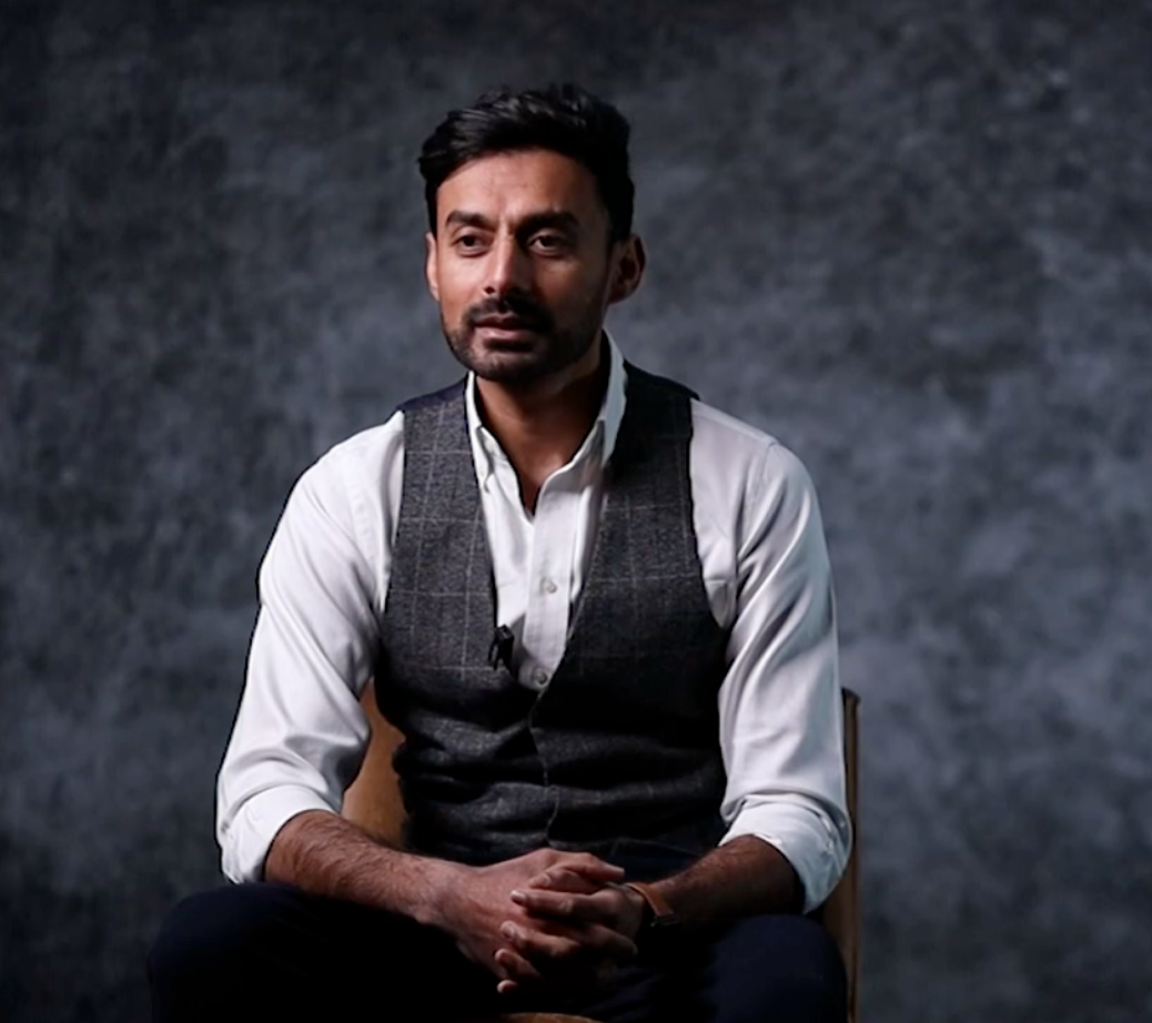 Studio portrait of a person in a white dress shirt and dark vest against a textured gray backdrop.