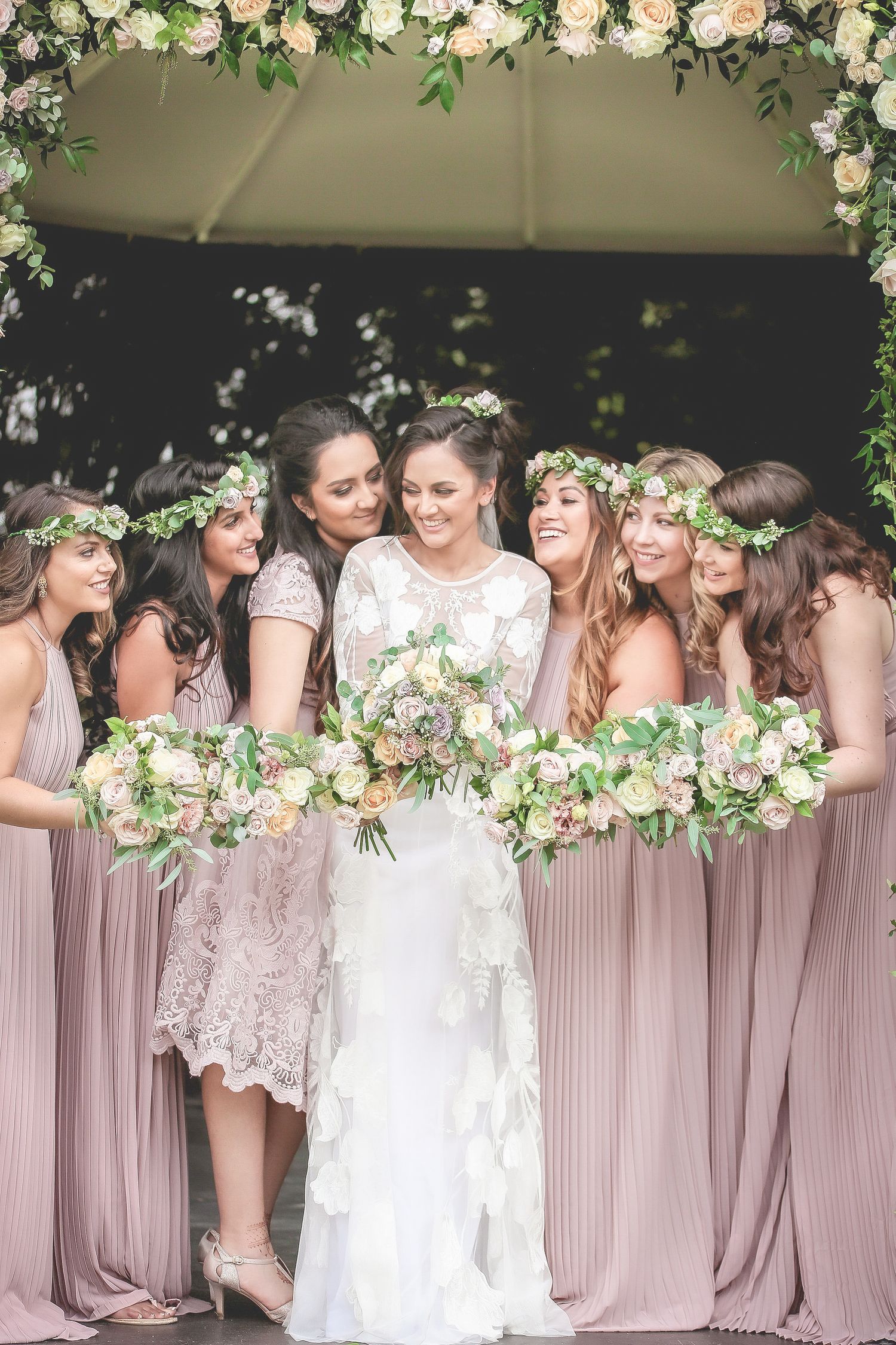 Bride in white lace gown surrounded by bridesmaids in dusty rose dresses holding floral bouquets under flower arch.