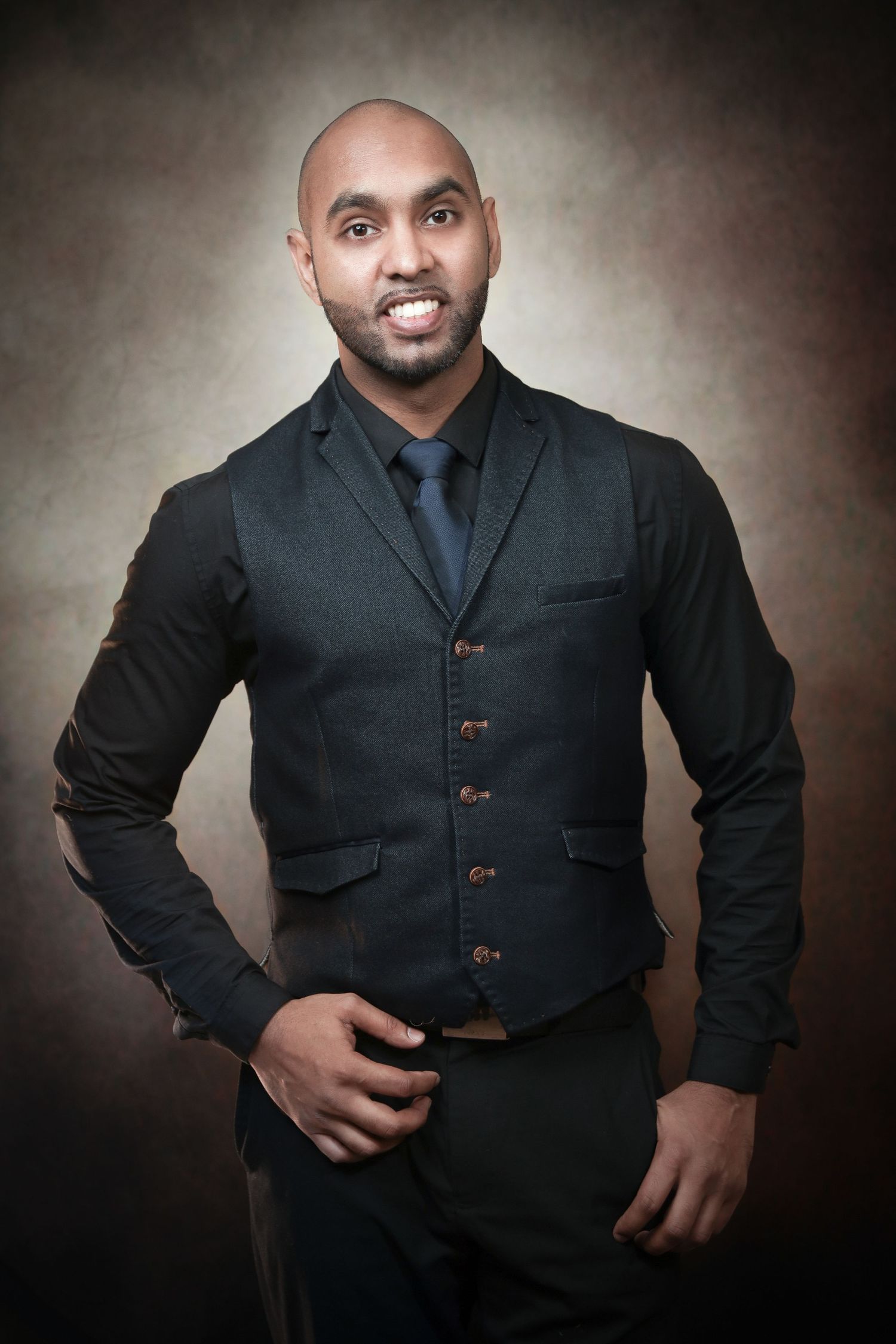 Bald man with beard wearing dark vest suit and tie posing confidently against brown studio background.