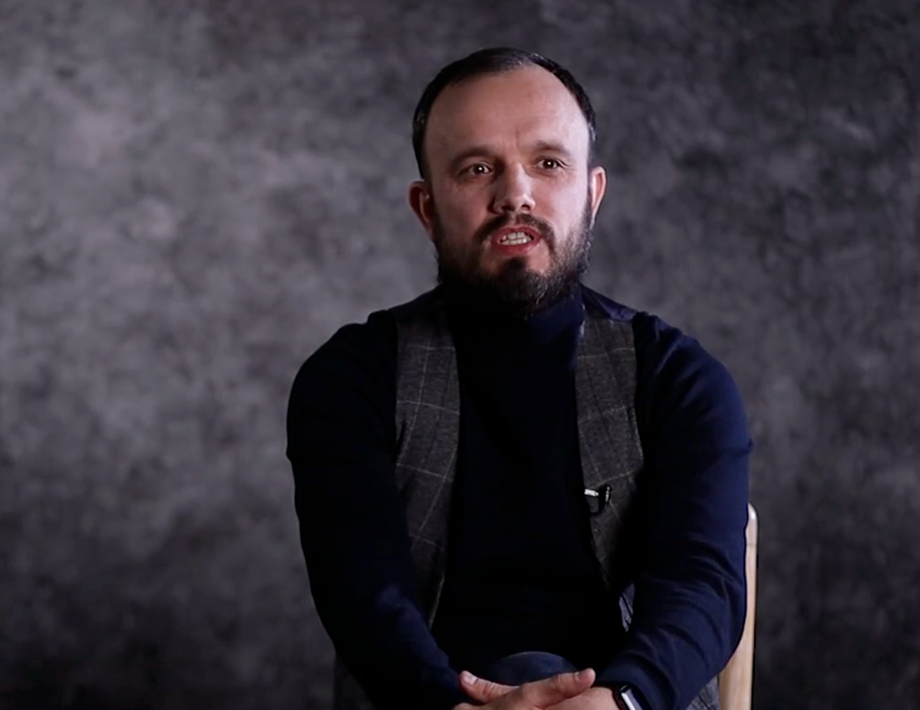 Professional studio portrait of person in navy blue sweater and black vest against dark gray textured backdrop.
