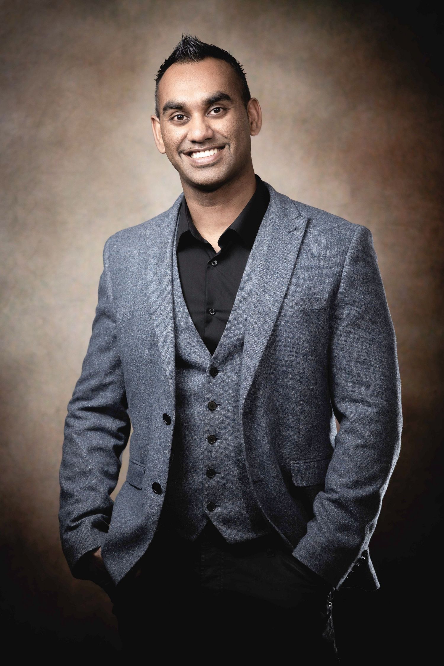 A smiling young man in a gray three-piece suit poses confidently with hands in pockets against a brown studio background.