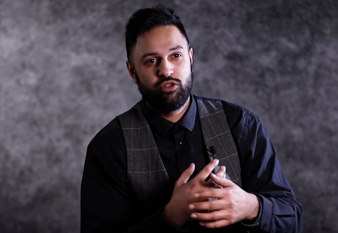 Professional portrait of someone wearing a dark checkered vest and navy shirt against a textured gray background.