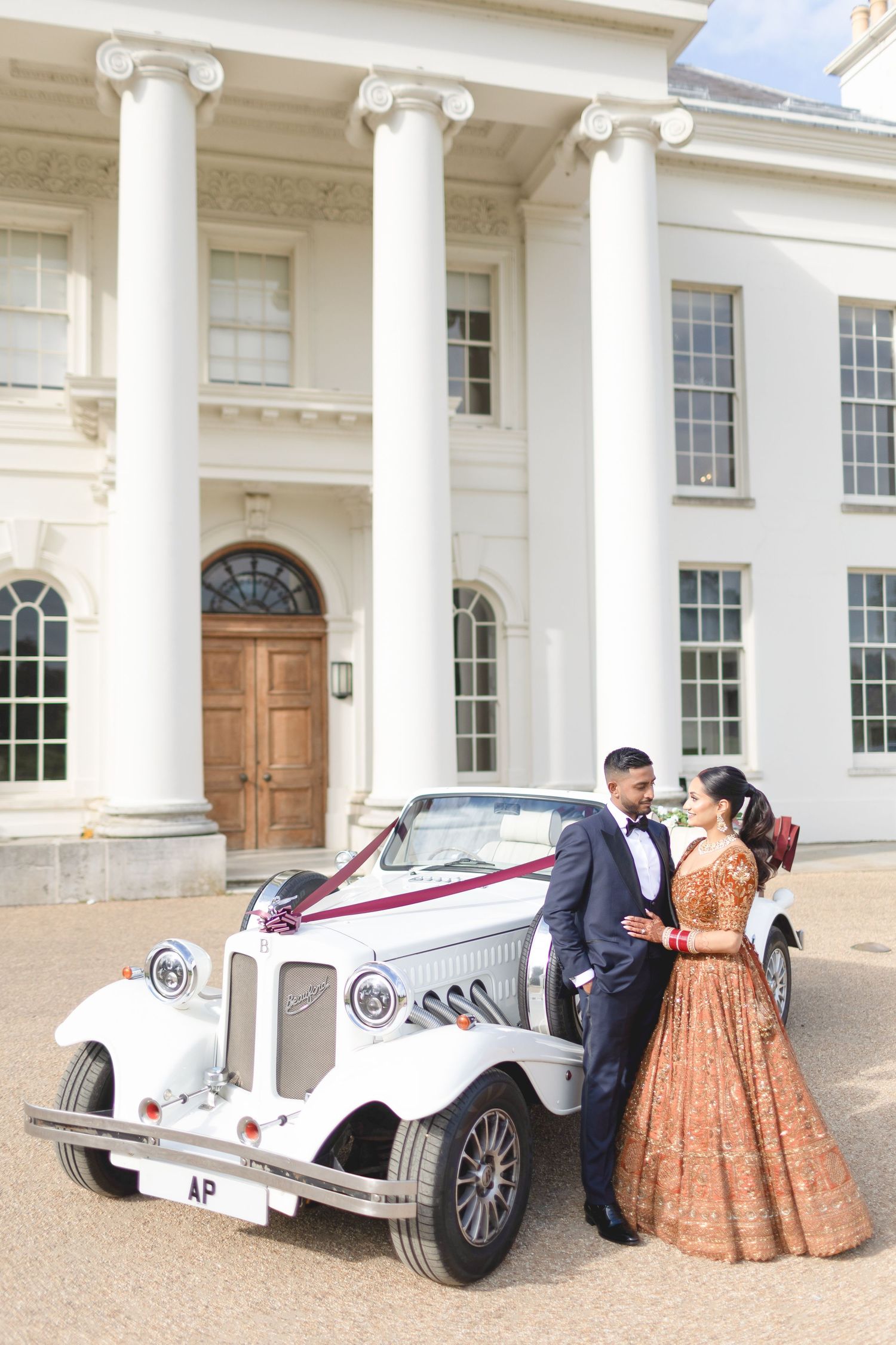 Bride and groom in elegant attire pose beside a vintage white car adorned with burgundy ribbons outside a grand neoclassical venue.