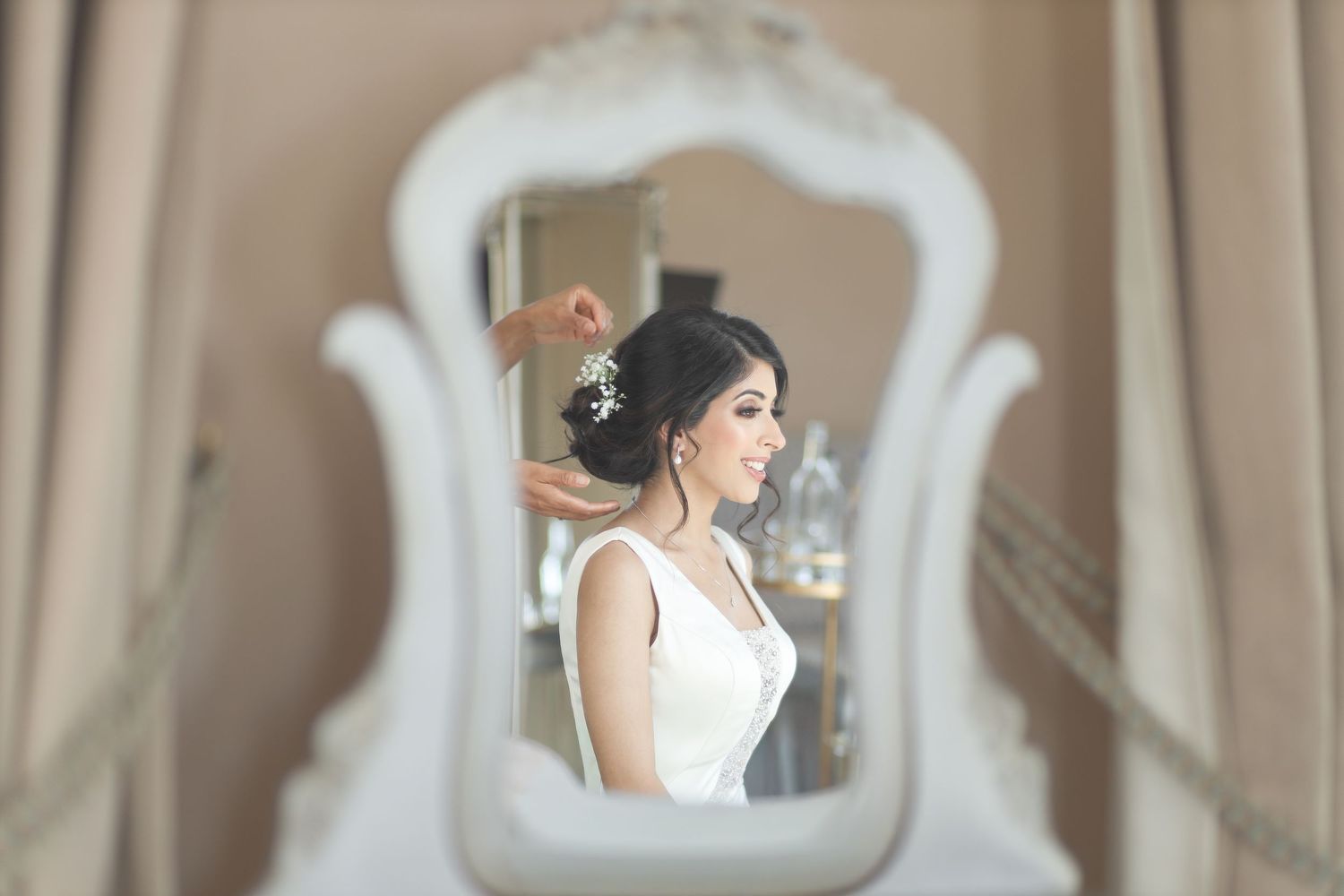 Bride in white dress getting hair styled, reflected in ornate white mirror during wedding preparations.