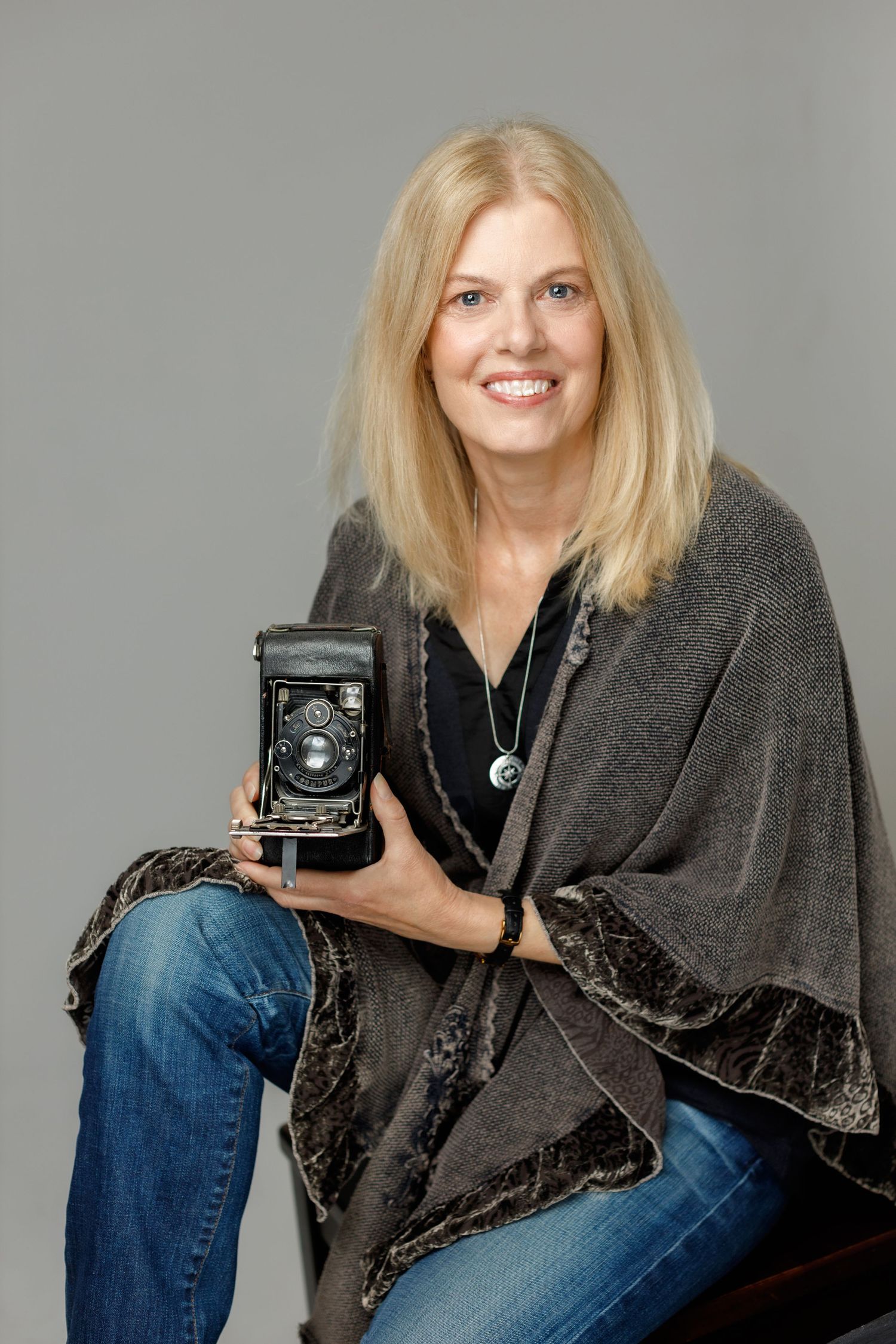 Professional photographer in blue jeans and black shawl holding vintage folding camera against gray background.
