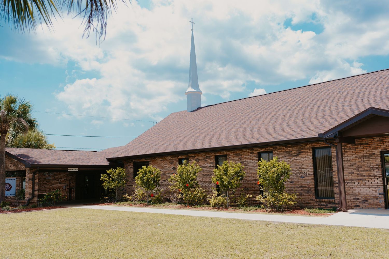Park Alliance Church; A panoramic view of a modern church building with a tall white spire against a blue sky with palm trees and clouds.