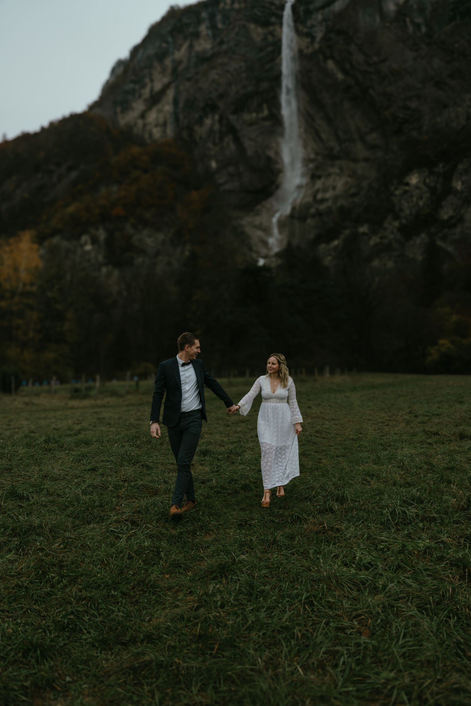 Couple walking hand in hand through meadow with dramatic waterfall backdrop.