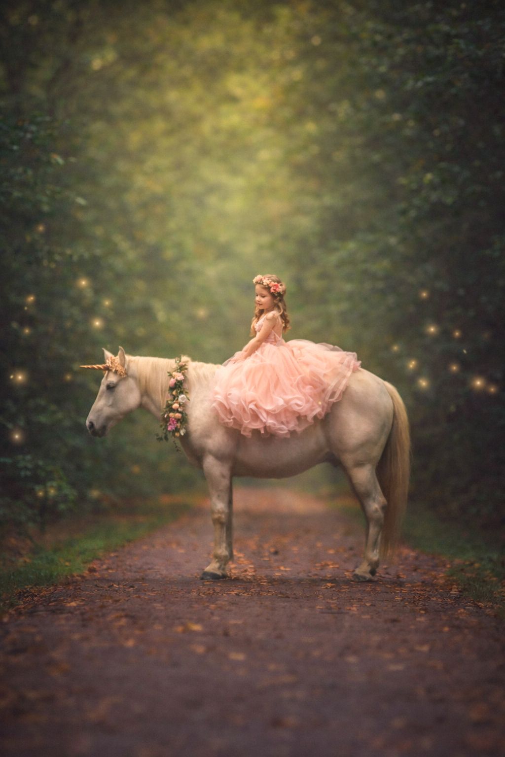 A young girl in a pink tulle dress sits atop a white unicorn on an enchanted forest path with fairy lights.