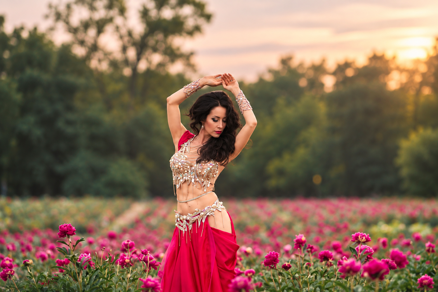 A woman in a red and gold belly dance costume poses gracefully in a vibrant flower field at sunset.