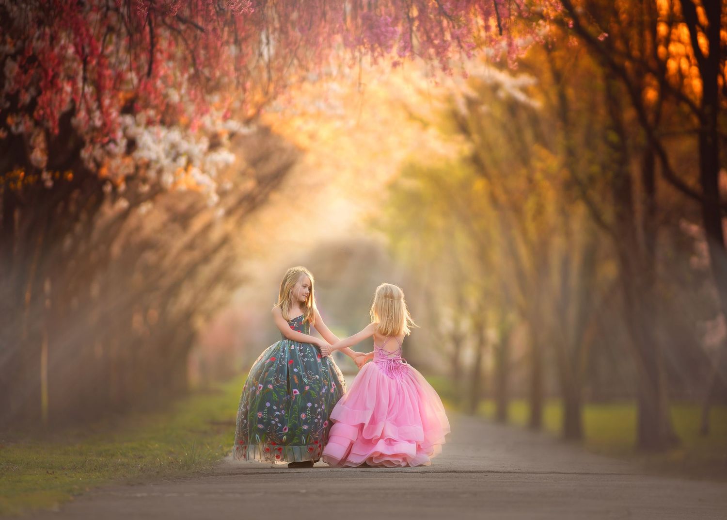 Two little girls in princess gowns dance on a tree-lined path bathed in golden sunlight.
