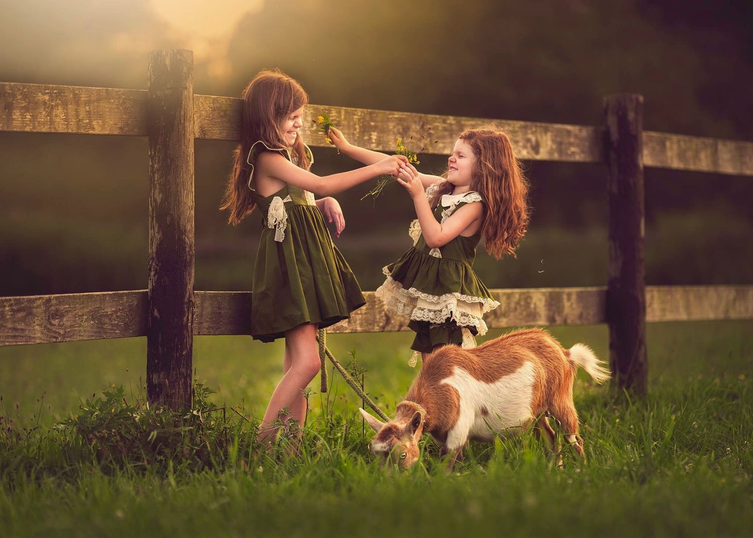 Two girls in green dresses share a moment by a wooden fence while holding a small goat on a sunny day.