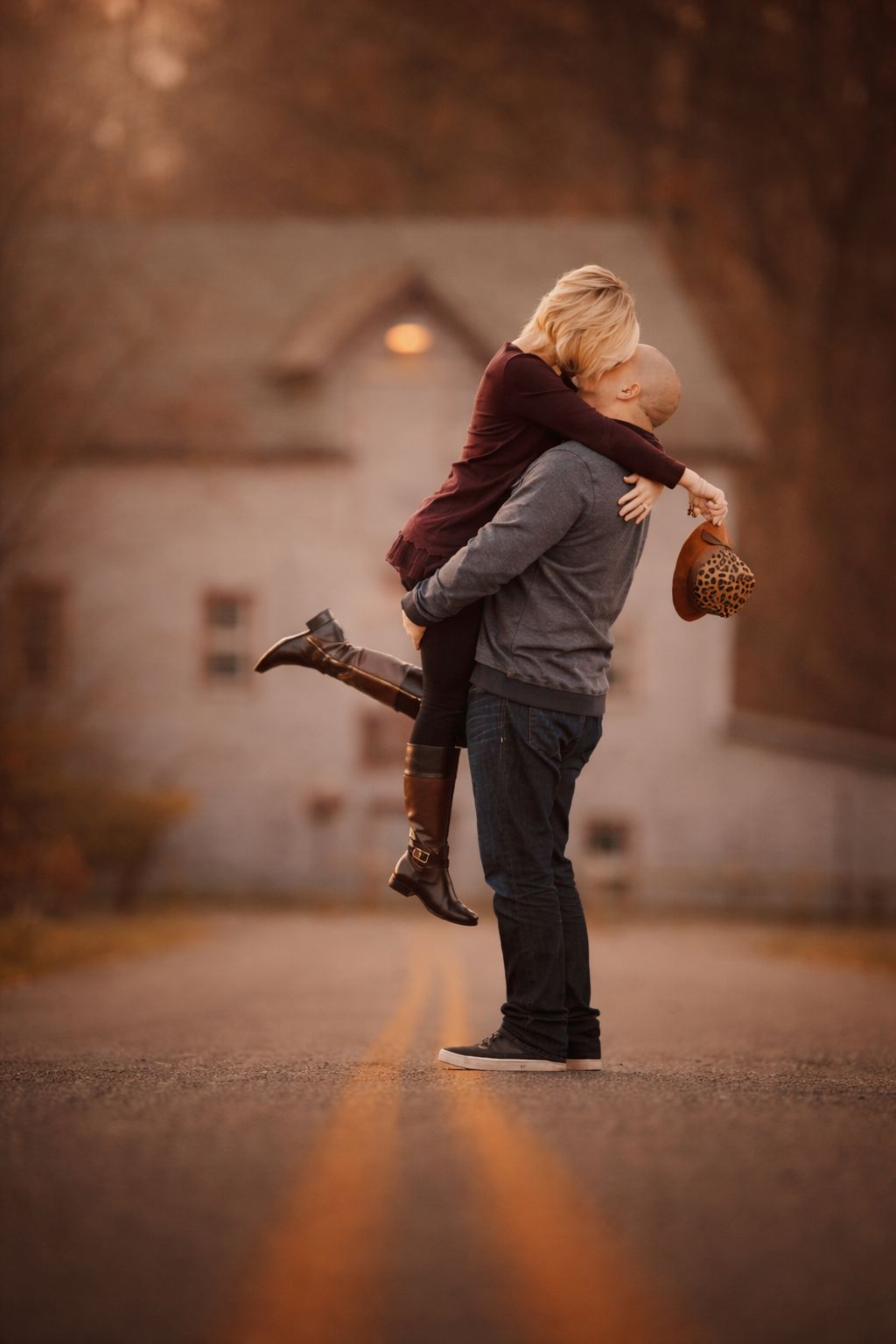 A couple shares a romantic embrace on a quiet road, man lifting woman in autumn setting with white farmhouse behind.