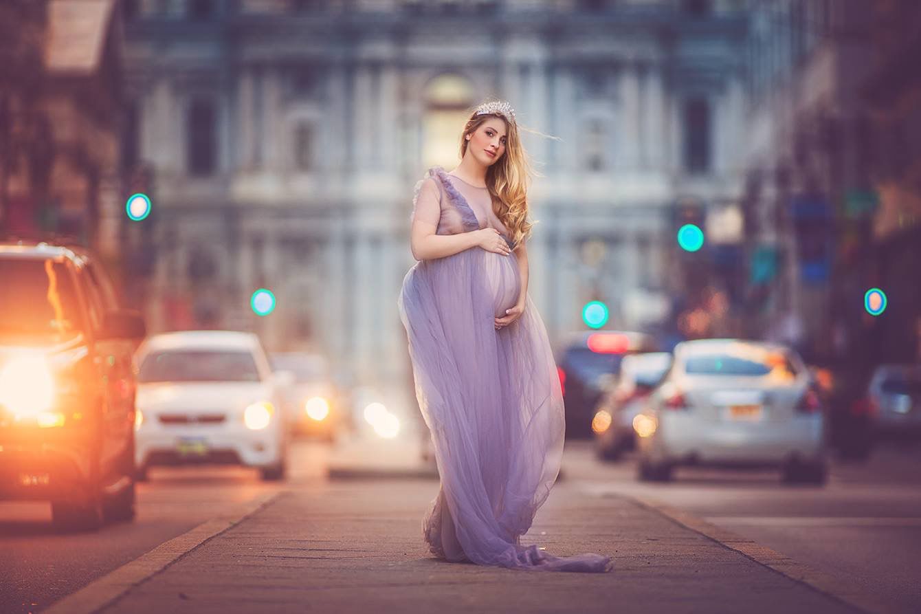 Pregnant woman in flowing lavender gown poses on city street at dusk with blurred traffic lights behind her.