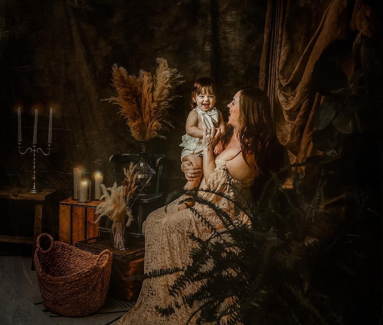 Mother in a lace gown holds her toddler beside candlelit decor, dried pampas grass and a wicker basket in a moody, vintage-style portrait.