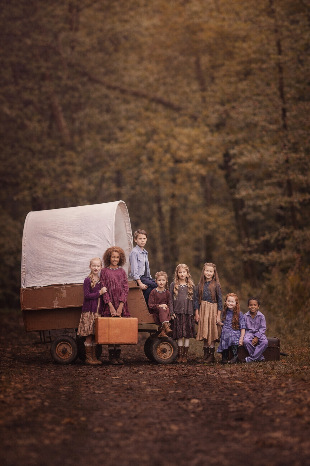 Children in vintage clothing pose with a covered wagon on an autumn forest path.