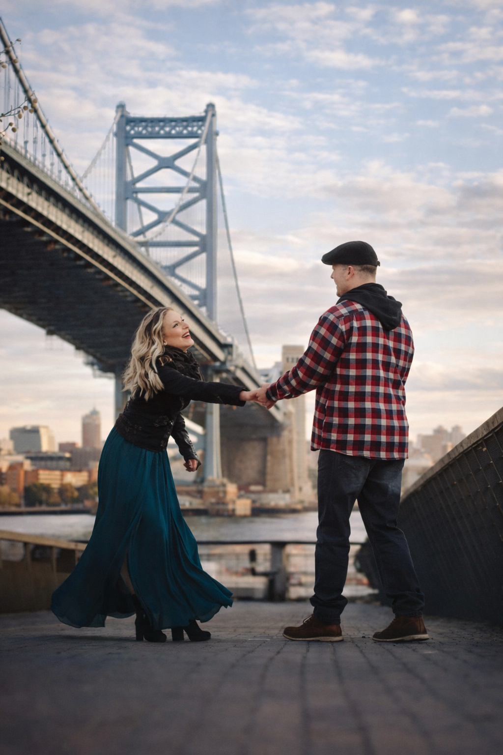 Couple holding hands and dancing on a waterfront boardwalk with the Benjamin Franklin Bridge and Philadelphia skyline behind them.
