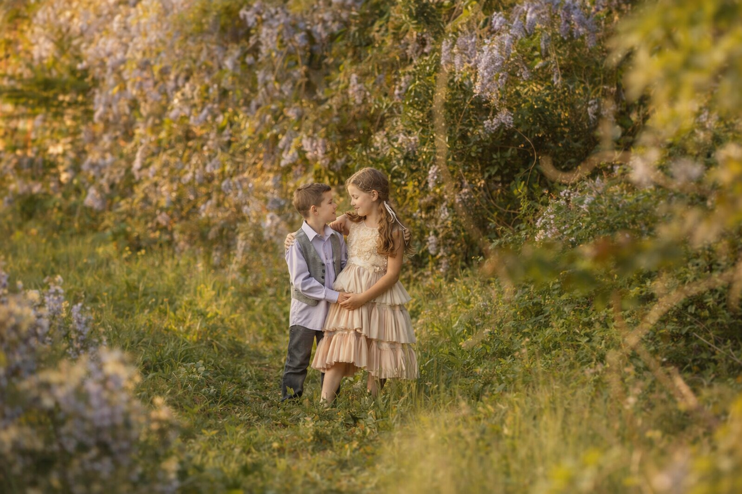 Two children in elegant attire embrace in a lush garden with blooming wisteria during golden hour.