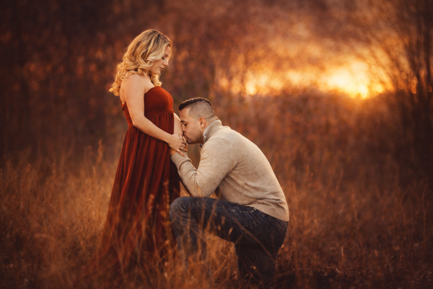Man kneels kissing pregnant womans belly in red gown during golden sunset maternity photo session.