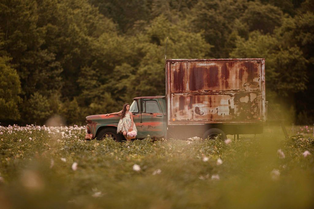 Woman standing beside a rusty vintage box truck in a blooming flower field with green trees in the background.