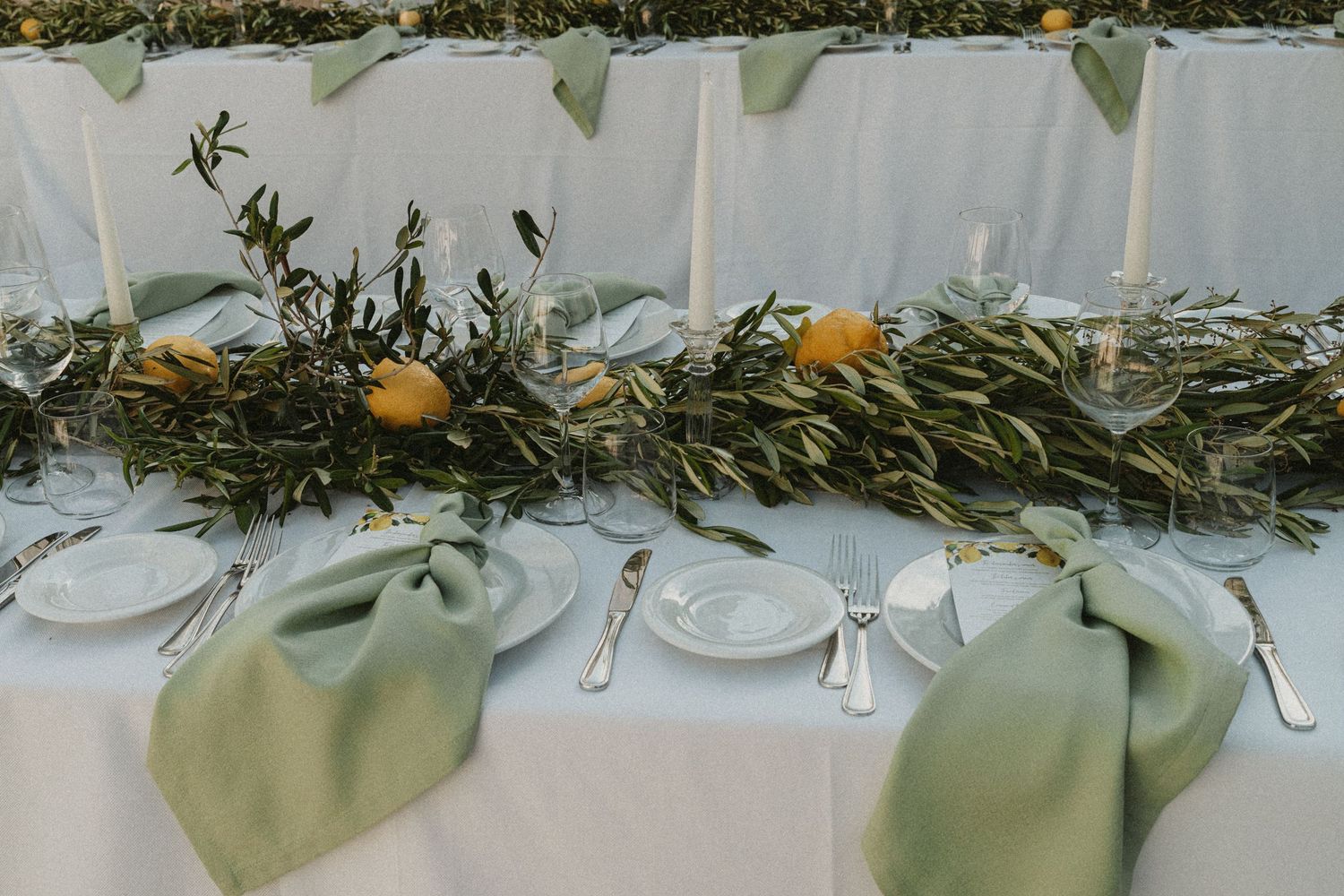A rustic wedding reception table decorated with olive branches, white plates, and natural greenery garlands.