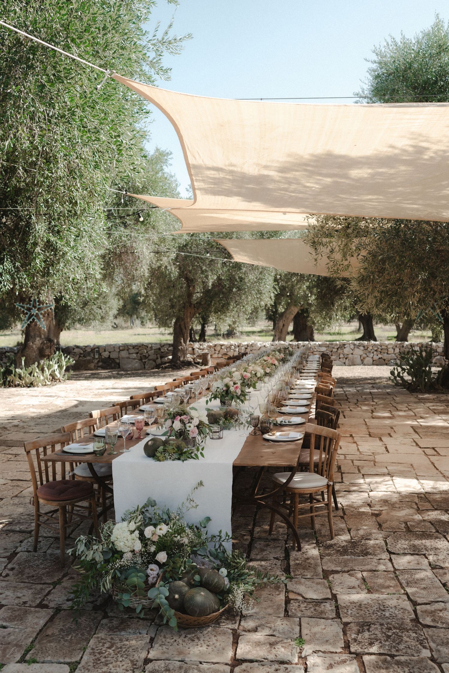 Rustic outdoor wedding reception setup with long tables under canvas shade sails and olive trees.