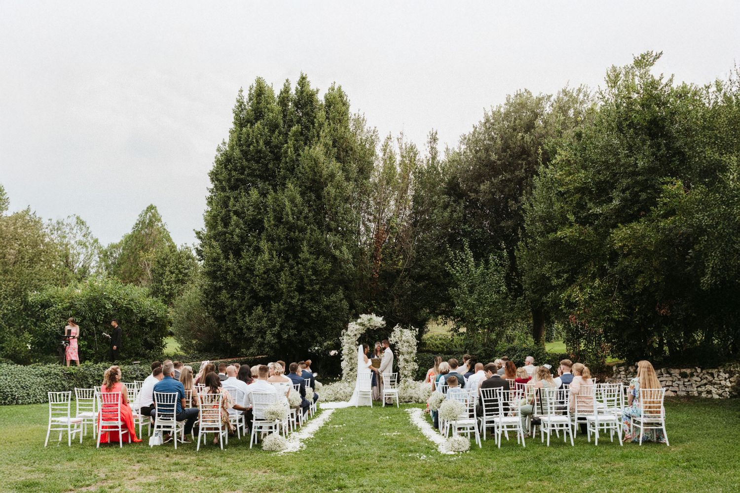 Outdoor wedding ceremony setup with white chairs arranged in rows on a green lawn surrounded by tall trees.