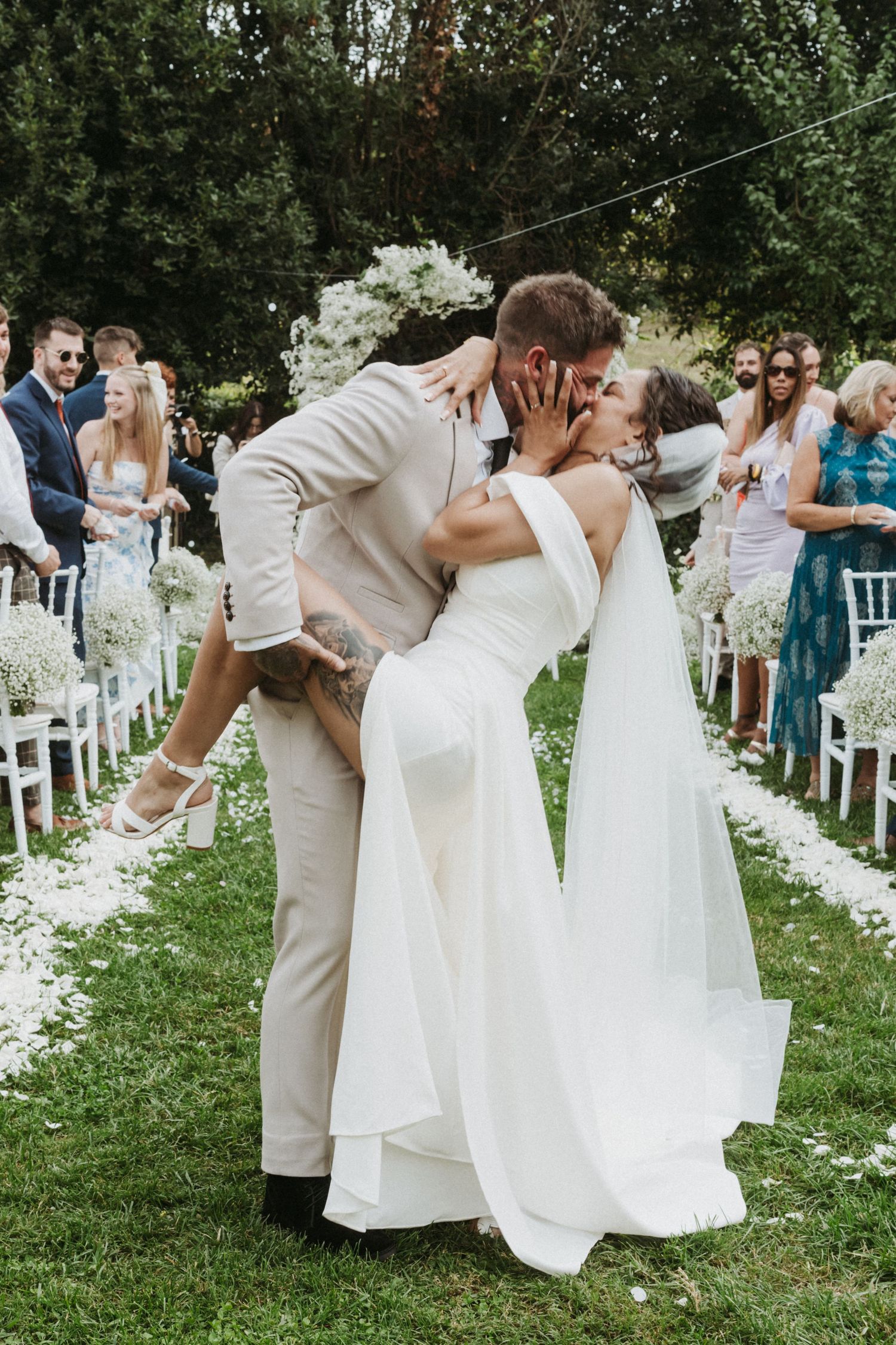Couples perform a coordinated dip dance move at an outdoor wedding ceremony surrounded by white chairs and greenery.