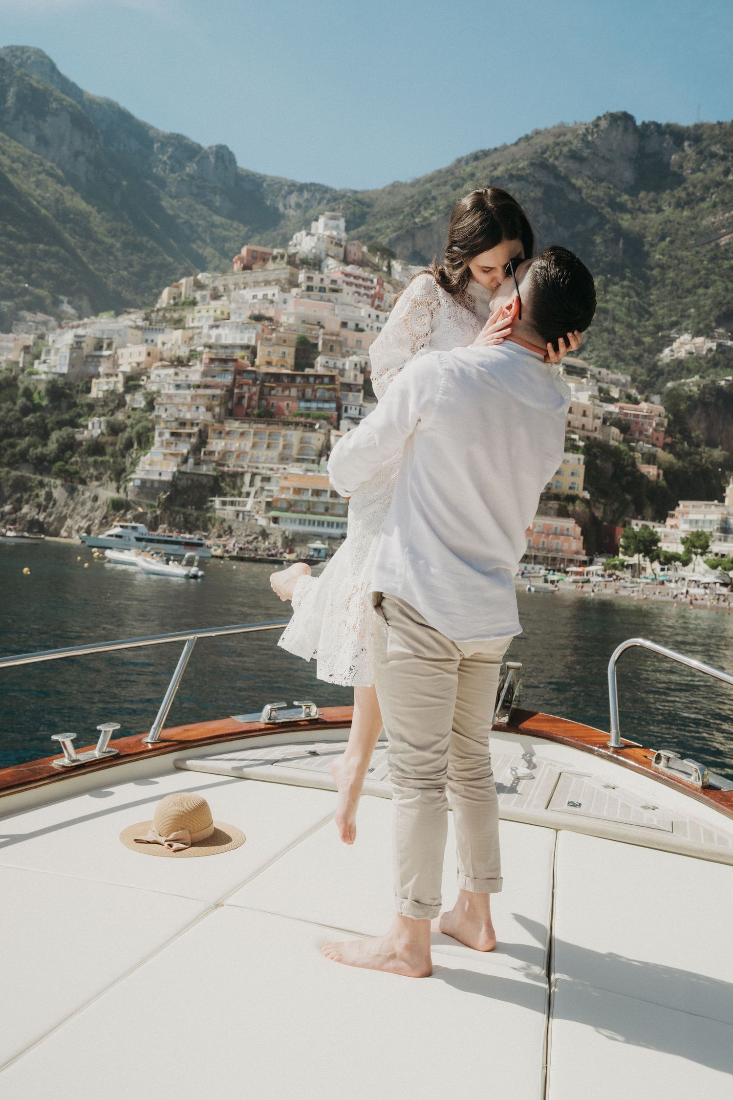 A couple in white outfits embracing on a boat deck overlooking the scenic Amalfi Coast and Mediterranean waters.