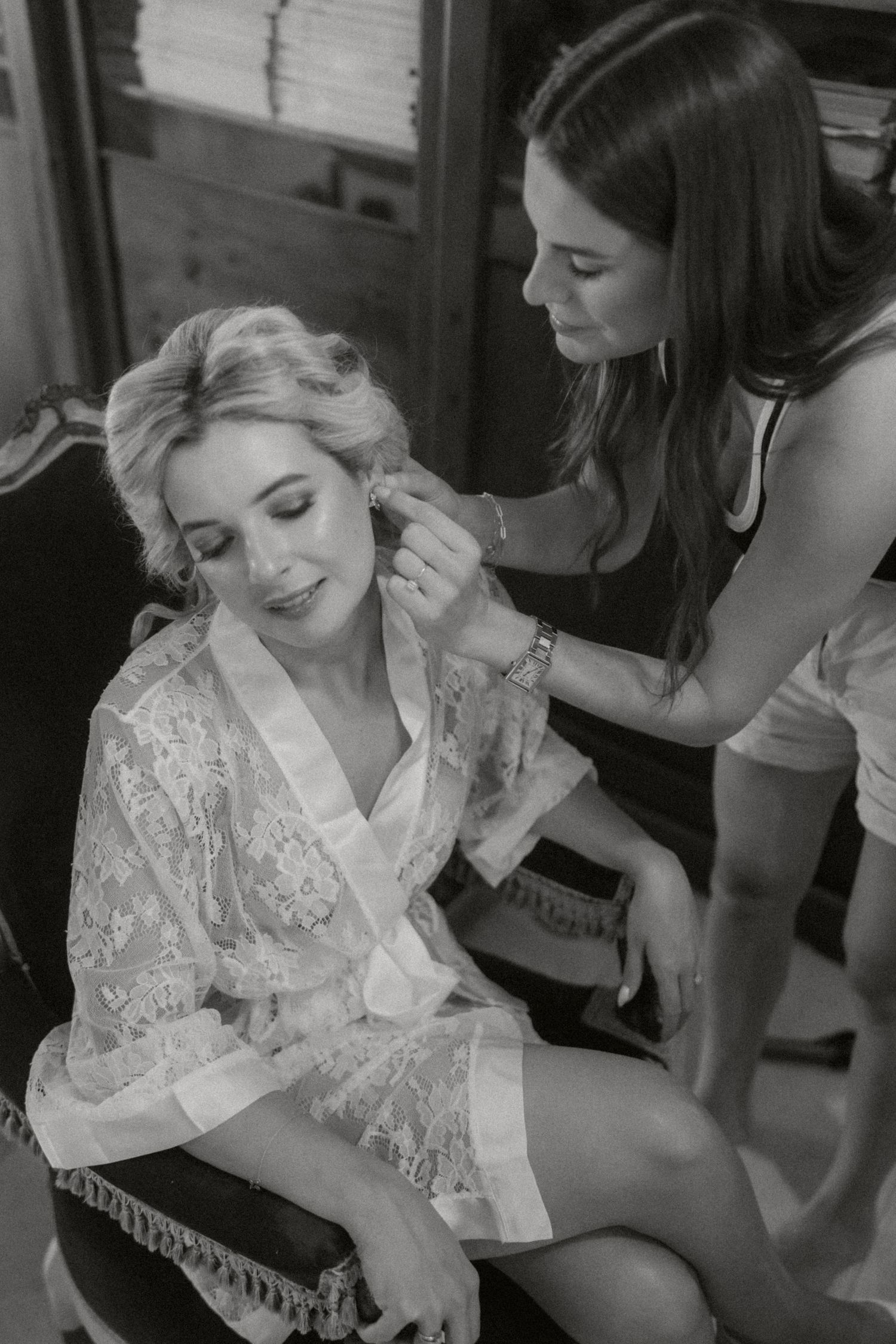 Black and white photo of a bride getting ready with her hair being styled while wearing a lace robe.