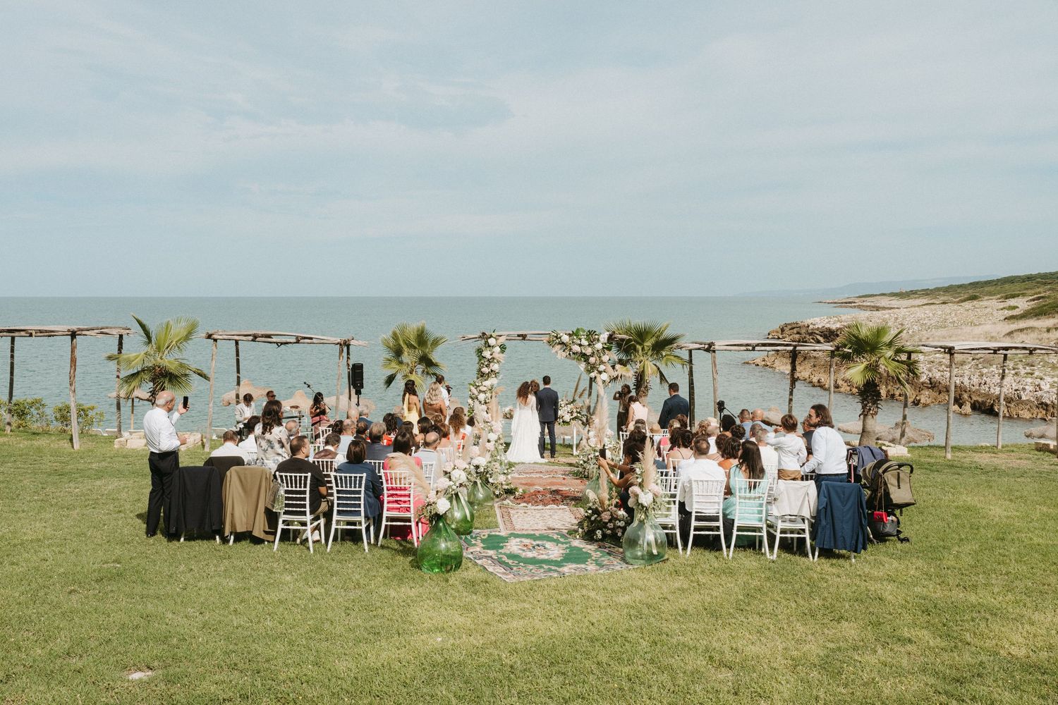 Panoramic view of an outdoor wedding reception setup with white chairs and tables on a grassy hilltop overlooking the ocean.