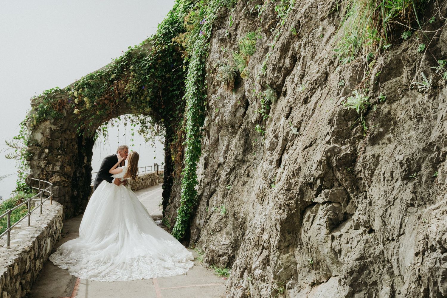 A couple in wedding attire embraces under an ivy-covered stone archway against a dramatic cliff wall.
