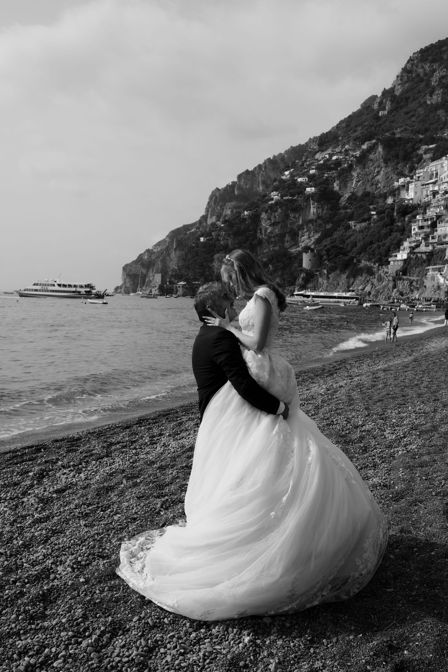 A couple in wedding attire sits together on a rocky beach with dramatic coastal cliffs in the background.