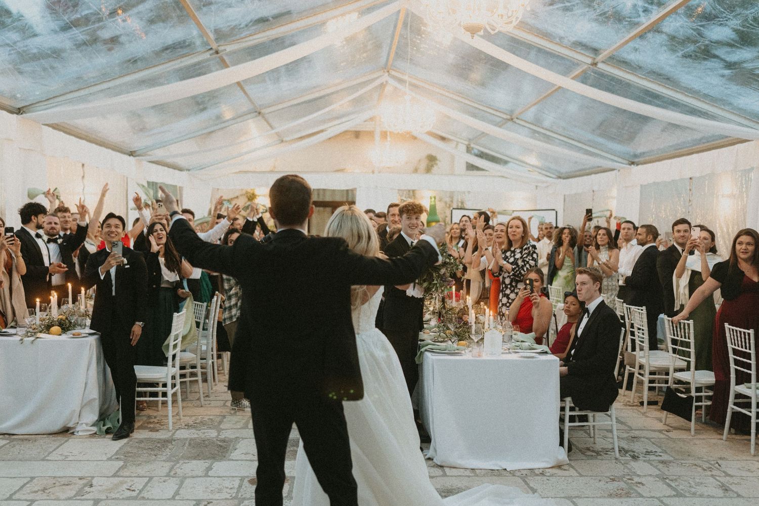 Wedding guests celebrate under a clear tent during an evening reception with string lights and white decor.