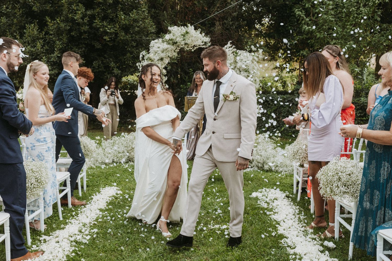 Newlyweds walking down aisle with white flower petals scattered on grass.