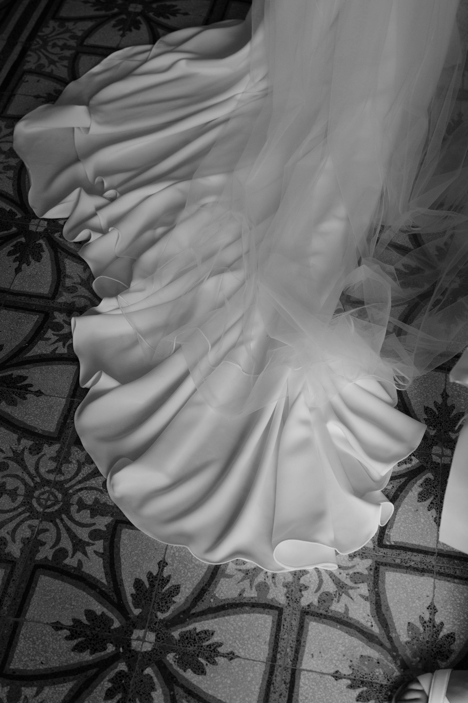 Close-up black and white detail shot of a white wedding dress hem flowing over an ornate patterned floor.