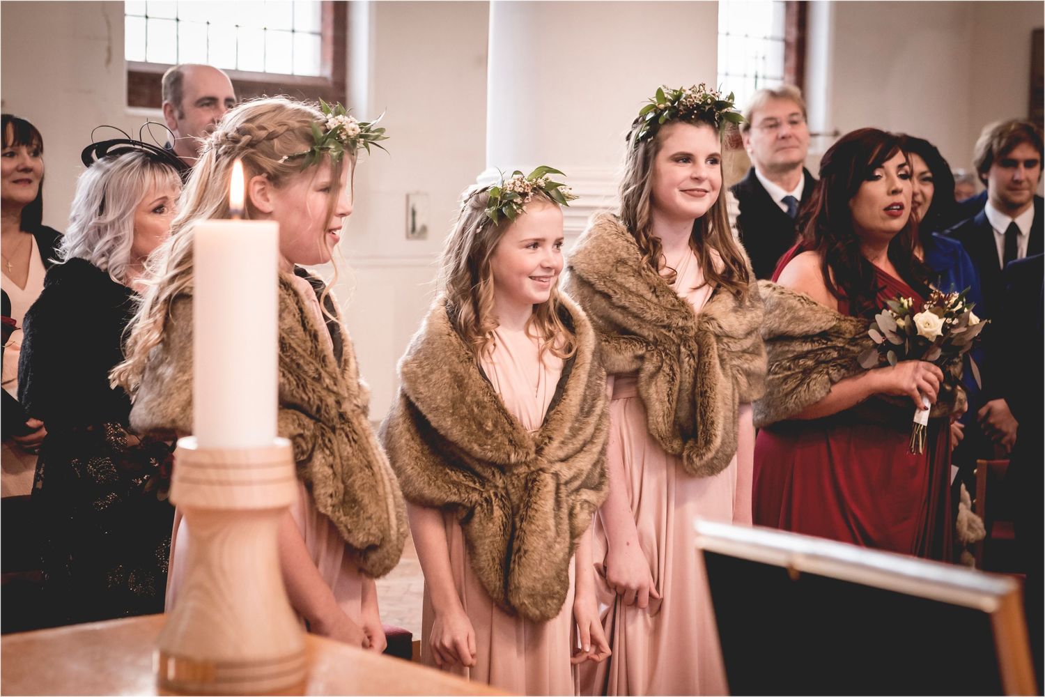 Bridesmaids in pink dresses and brown fur stoles stand together during a winter wedding ceremony in a church.