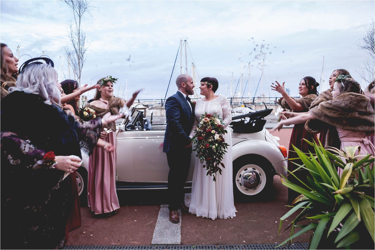 Wedding guests shower confetti on newlyweds next to vintage car at seaside venue on cloudy day.