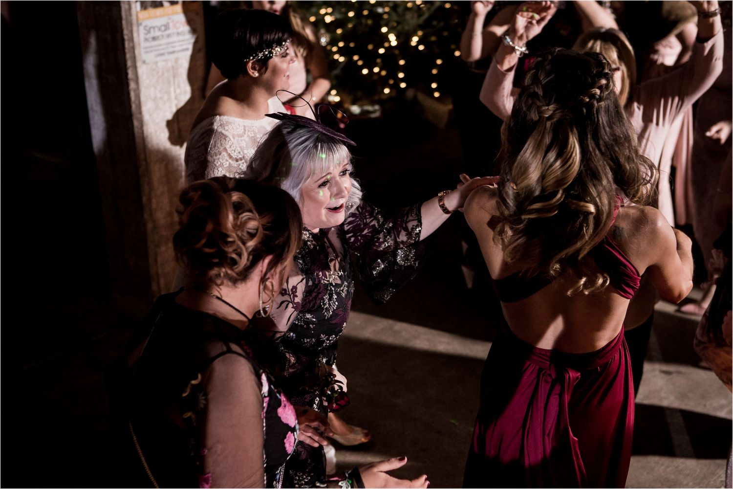 People dancing and celebrating at a dimly lit party with Christmas lights visible in the background.