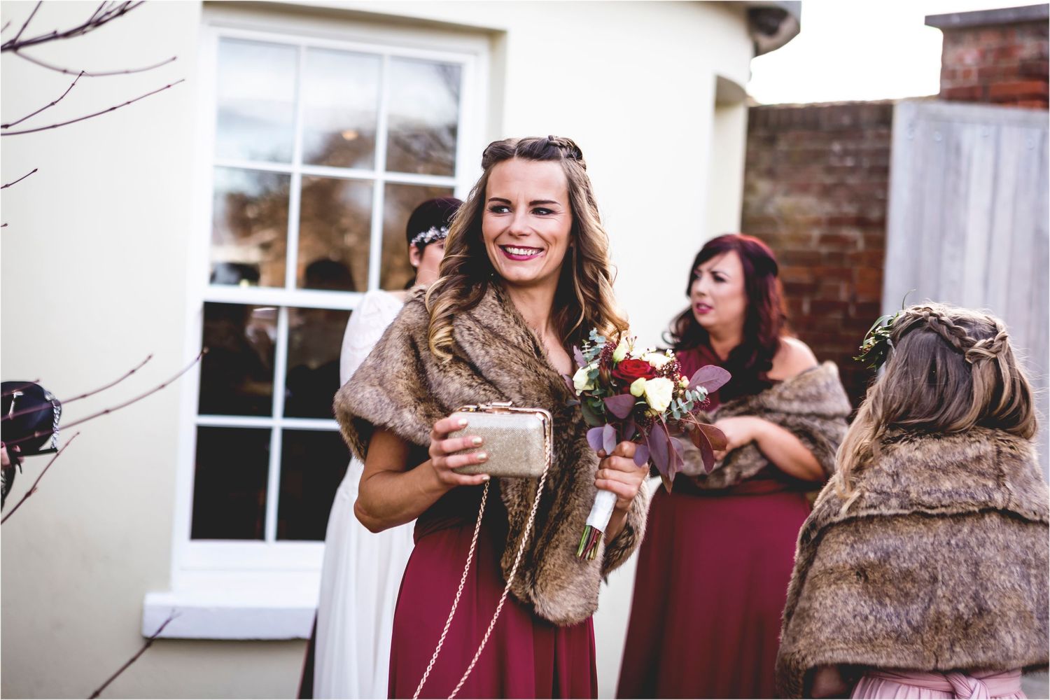 Wedding guests in burgundy dresses and fur shawls gather outside during a winter celebration.