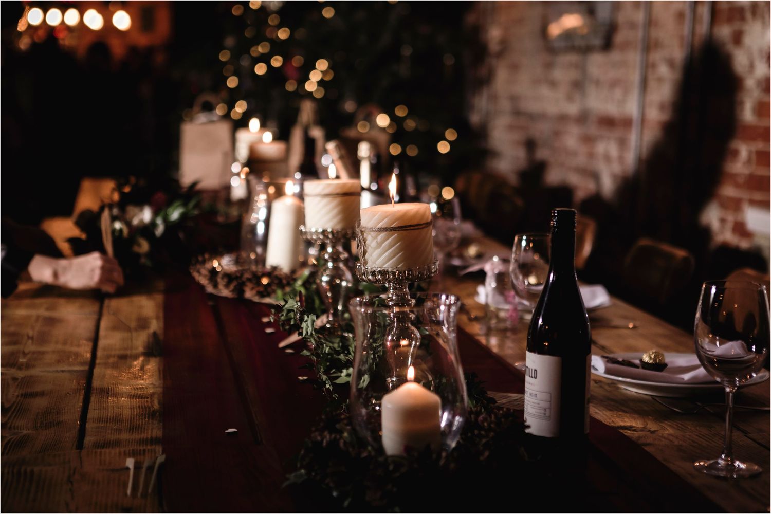 Rustic holiday table setting with lit candles, greenery, wine bottles and glassware on wooden surface in dim lighting.