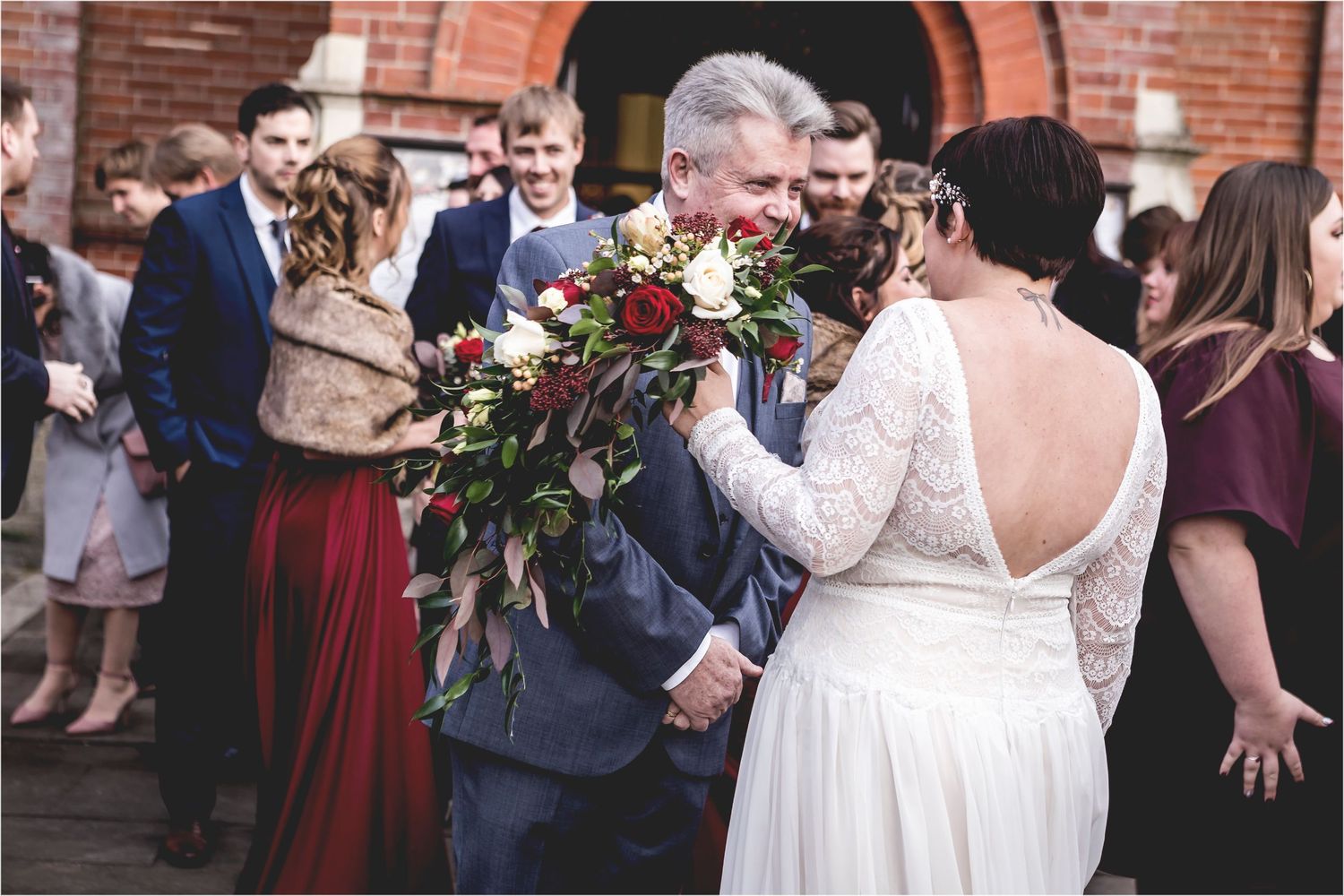 A couple shares a tender moment at their wedding celebration outside a brick church as guests look on in celebration.