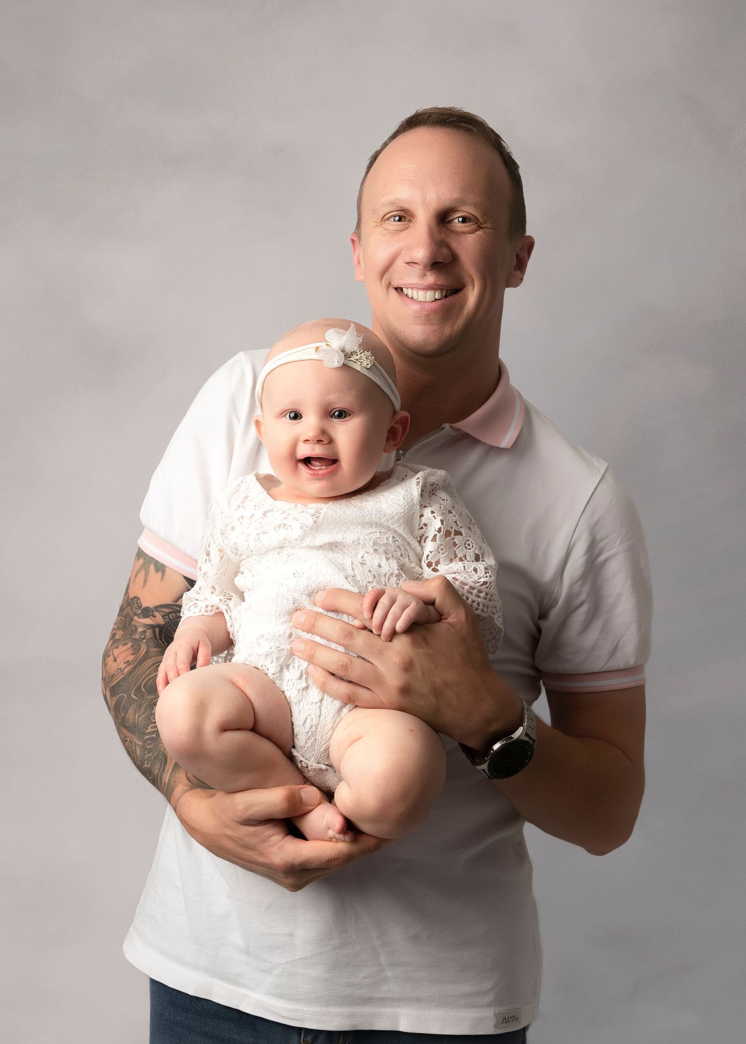 Smiling man with tattoos holds a happy baby girl in a white lace outfit and floral headband in a studio portrait.
