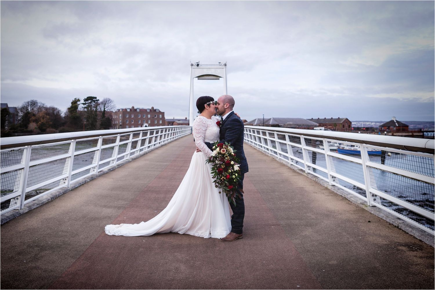 A romantic wedding couple shares a kiss on a white bridge overlooking the water under moody cloudy skies.