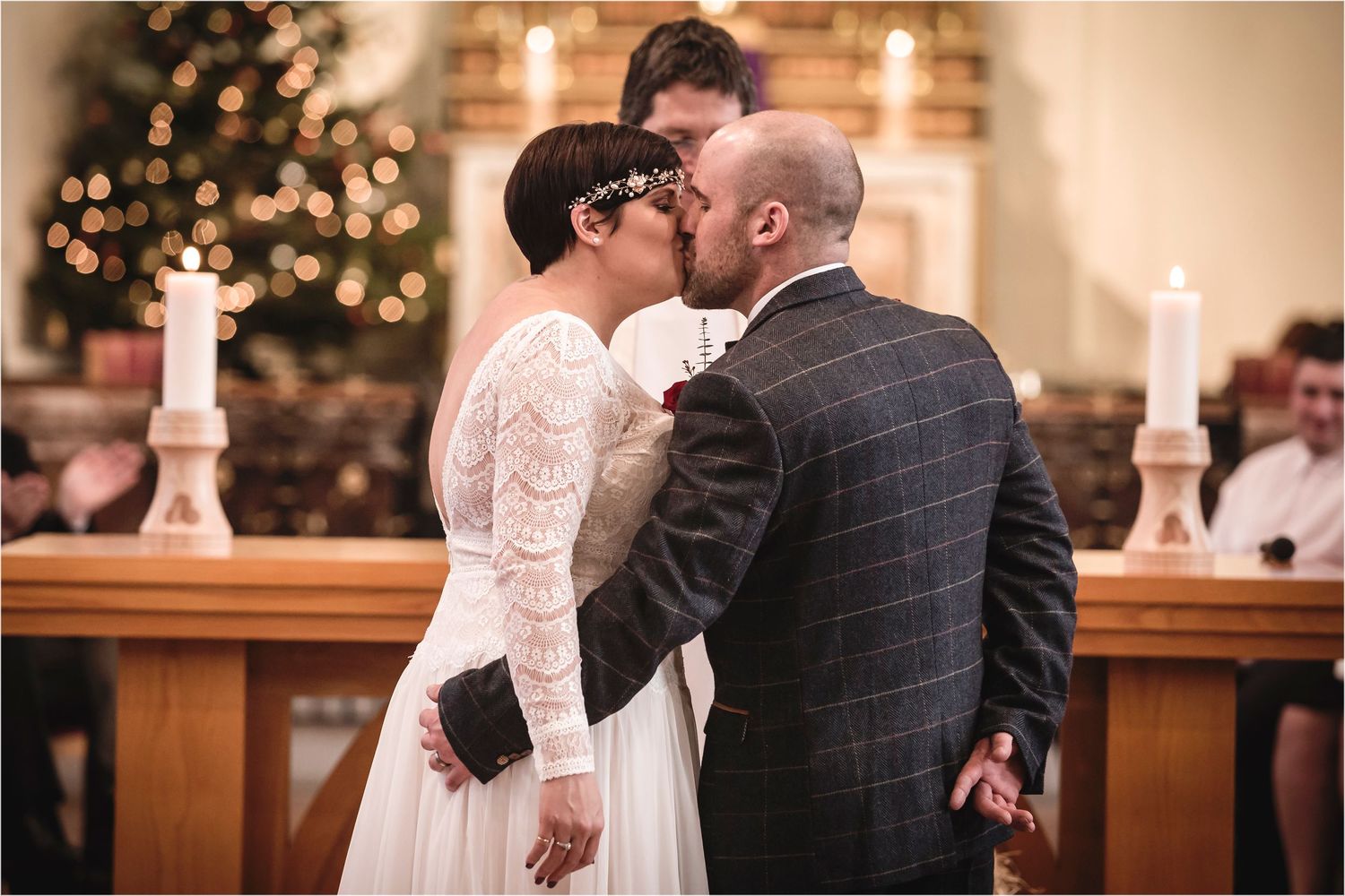 A romantic wedding ceremony kiss at the altar with candlelight and Christmas tree lights glowing in the background.