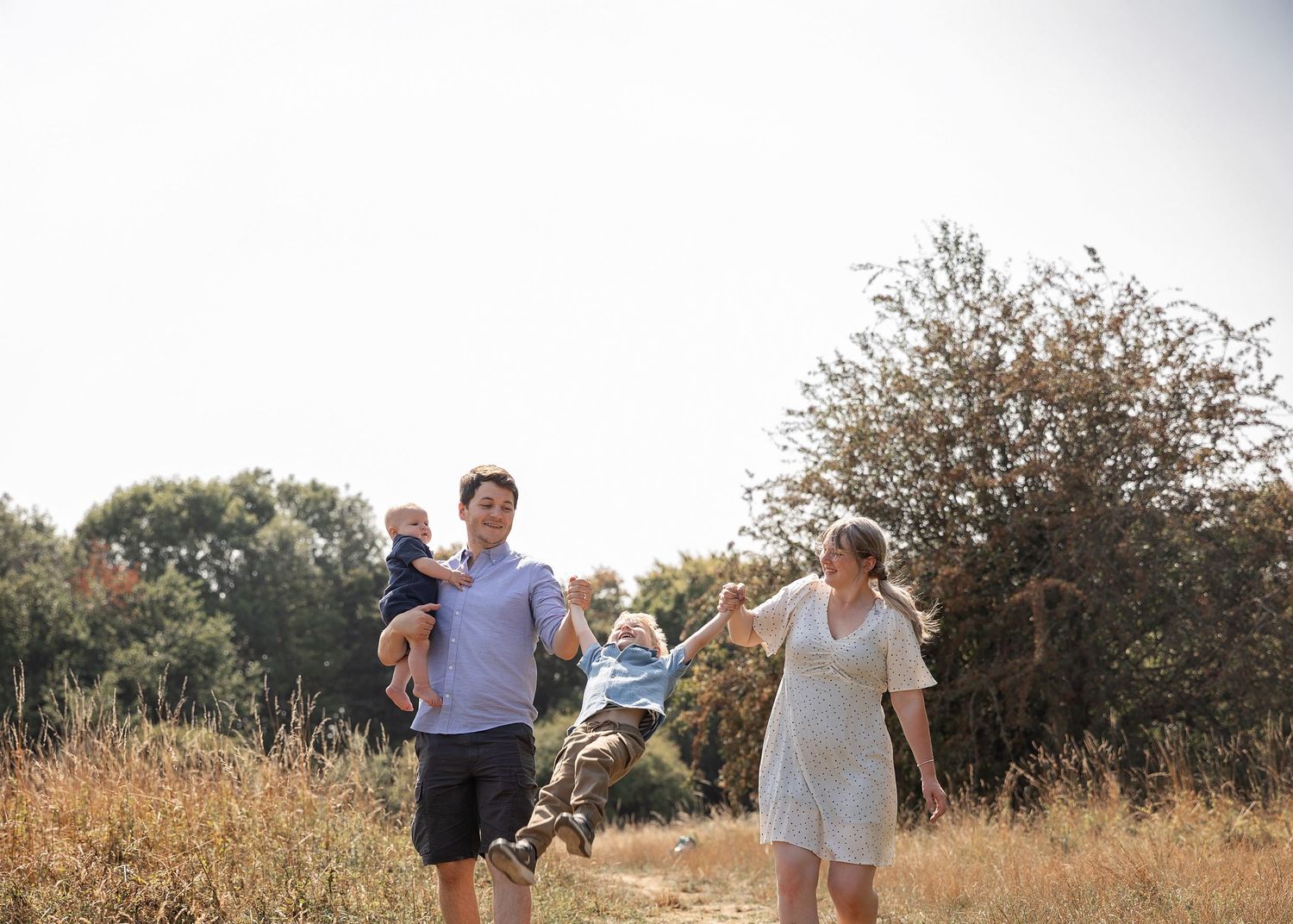 Family walking outdoors on a sunny day, parents carrying toddler through a golden field with trees in background.