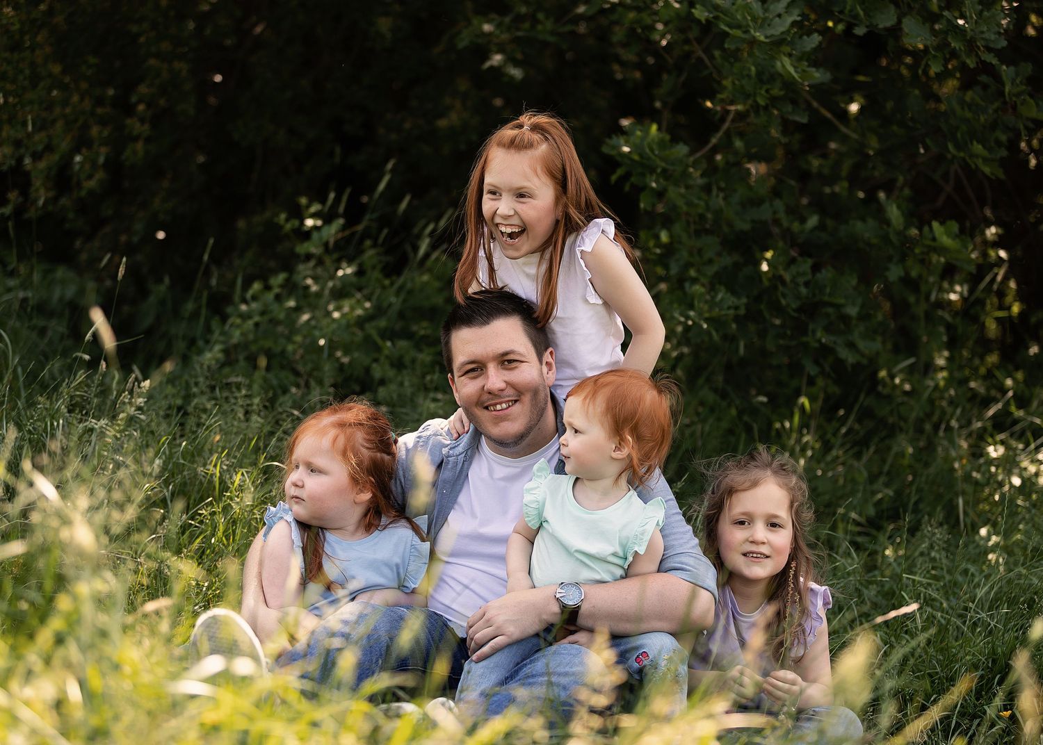 A father sits in a lush green meadow smiling with his four young redheaded children gathered around him.