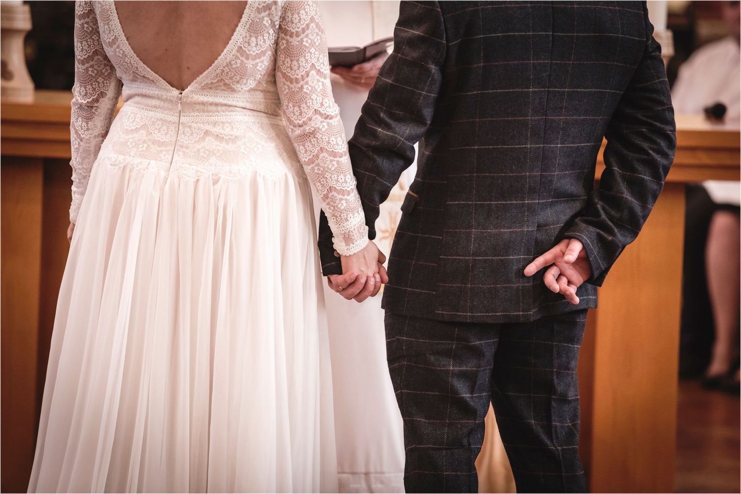 A couple holds hands at their wedding ceremony, with the bride in a lace-sleeved white gown and groom in a checked suit.