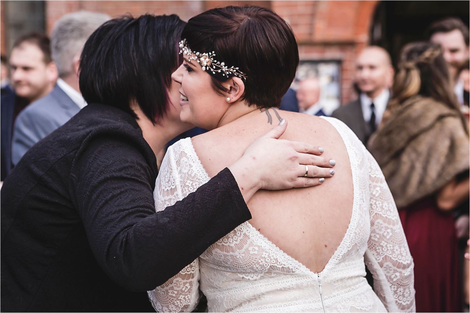 Intimate embrace between wedding couple wearing white lace dress and black suit during outdoor ceremony gathering.