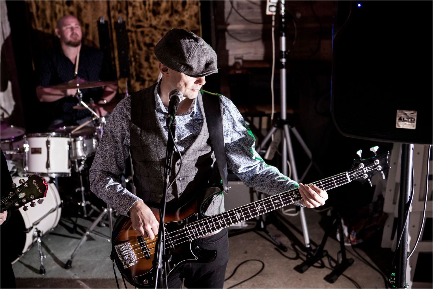 A musician performs on stage playing bass guitar while wearing a vest and hat during a live performance.