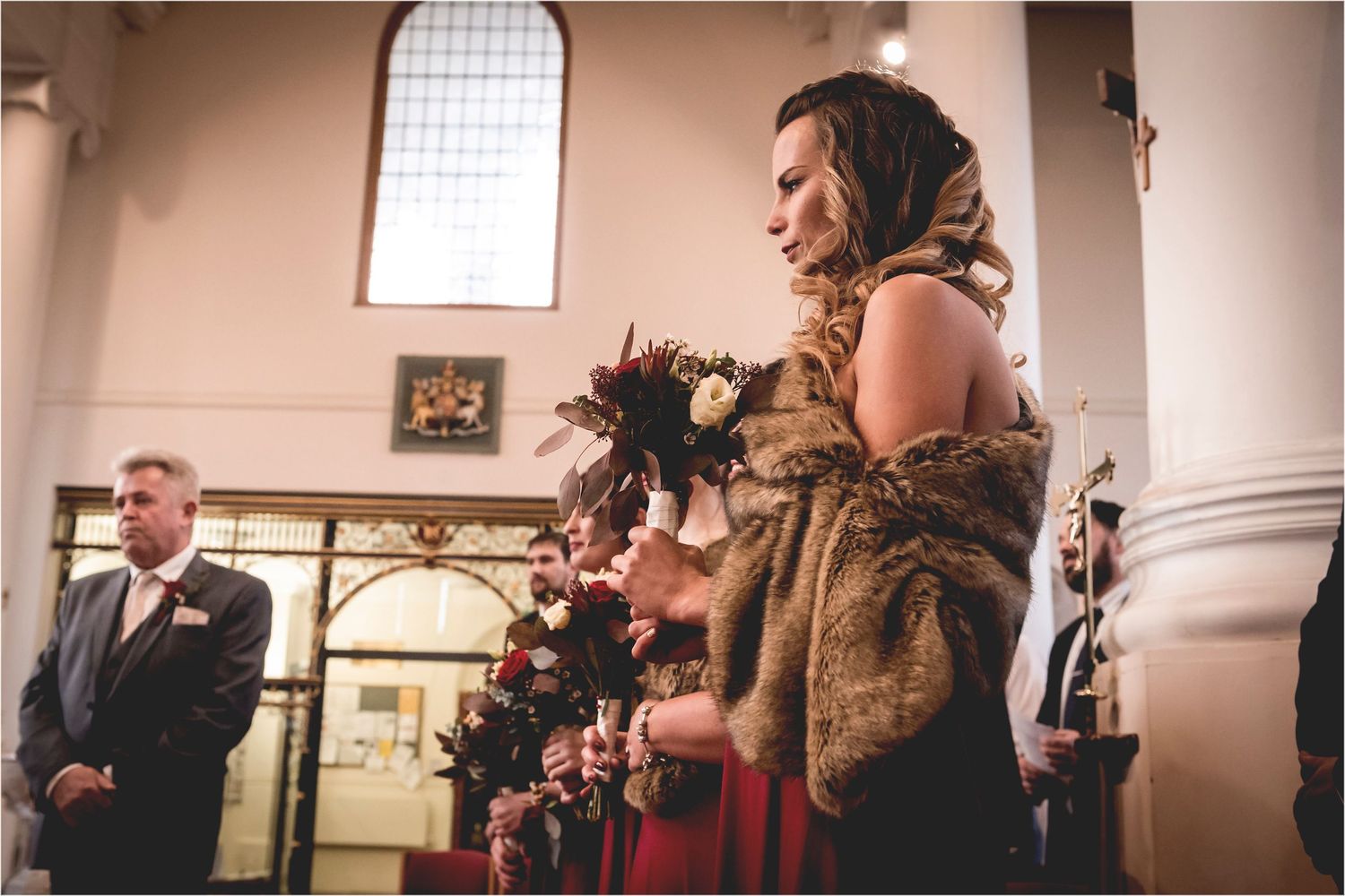 A bridal party stands in line at a traditional church wedding ceremony with elegant fur stoles and red dresses.